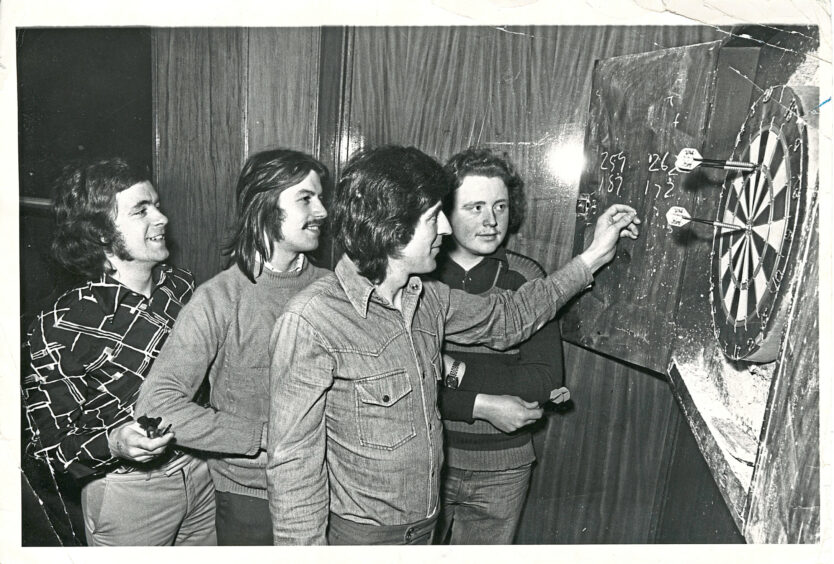 Four men stand in front of a darts board