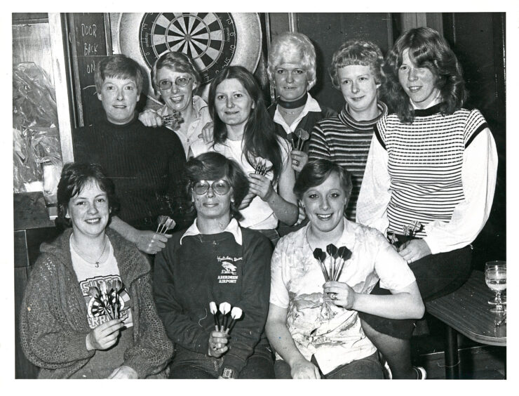 A woman's Aberdeen darts club gathers in front of a darts board.