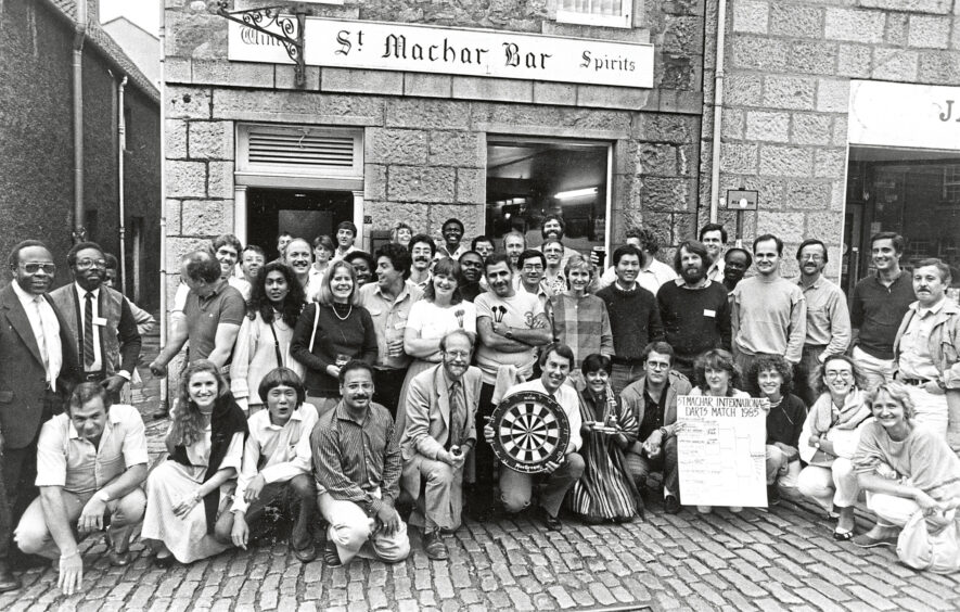 A large group of people stand in front of the St Machar Bar in Aberdeen. A person crouching at the front of the crowd holds up a dart board and another displays a scoreboard.
