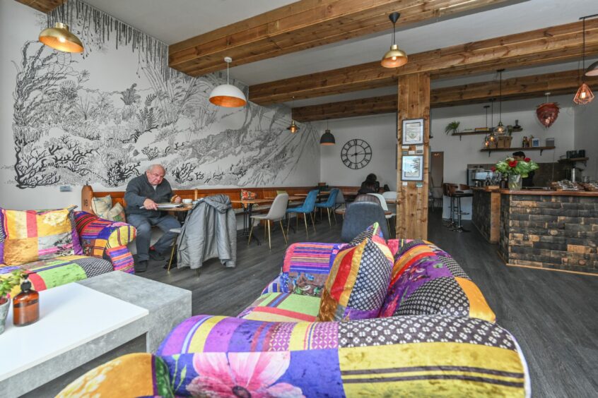 Colourful couches sit to the front of Utopia Cafe in Inverness, with customers scattered throughout the seating area, which has a feature wall and wood beams across the ceiling and room. 
