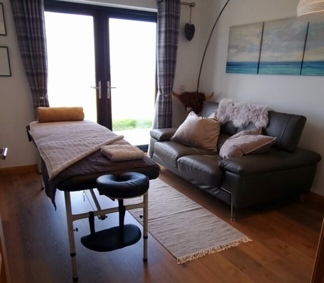 A room with a massage bed, sofa, and french windows looking out over Uist.