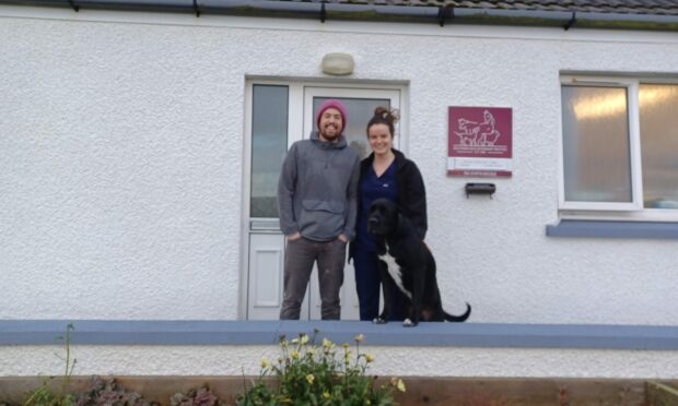 A man, a woman, and a large black dog pose outside Southern Isles Veterinary Practice