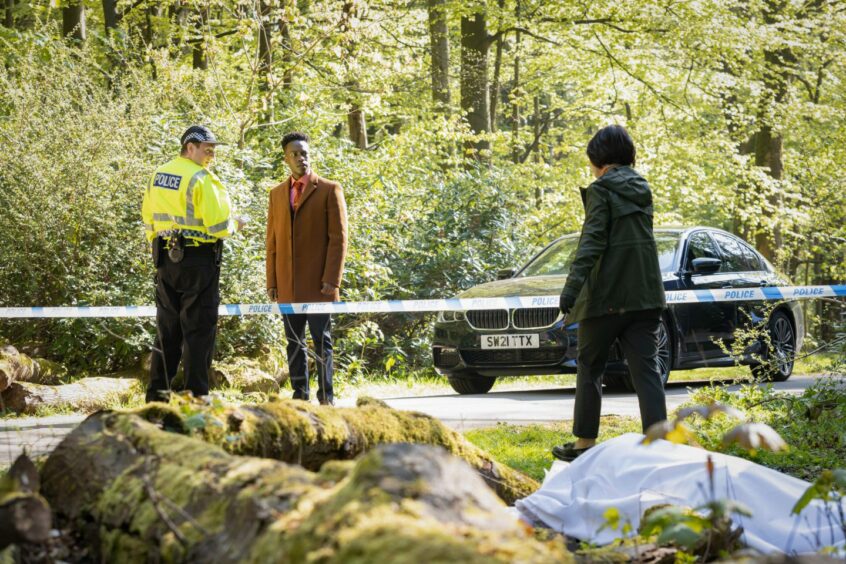 A still of Hannah Donaldson, Romario Simpson and a police officer extra from Granite Harbour with a body under a white sheet in the woods, taped off by police.
