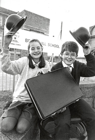 A boy and girl sat in front of Broomhill School, holding bowler hats in the air and holding a briefcase between them