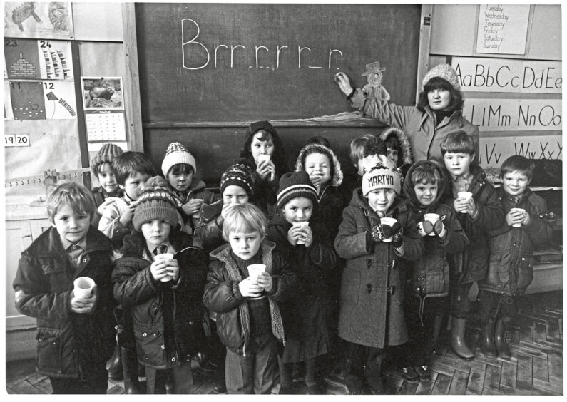 A class of young Broomhill School pupils with cups of hot orange standing in front of the blackboard. Their teacher points to the word "Brrrrrr" written on the board.