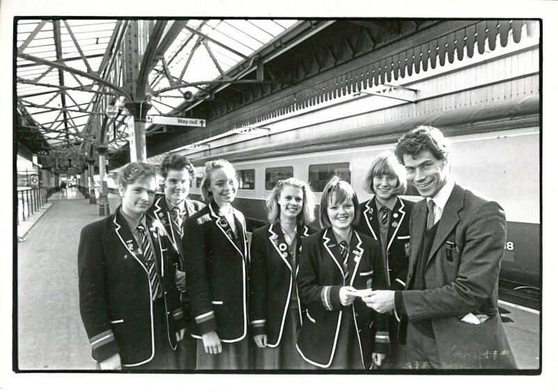 Six girls smile at the camera while one is handed a train ticket by a man