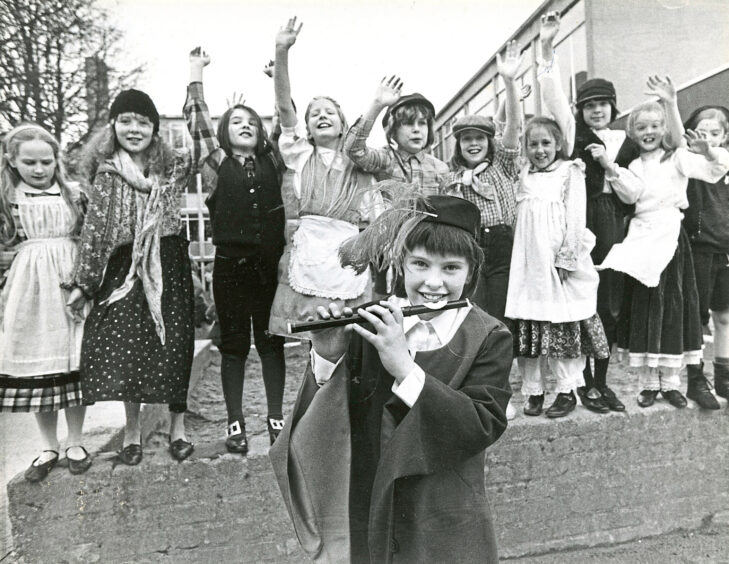 A girl with a flute smiles at the camera while a group of girls stand on the wall behind her with their hands in the air.