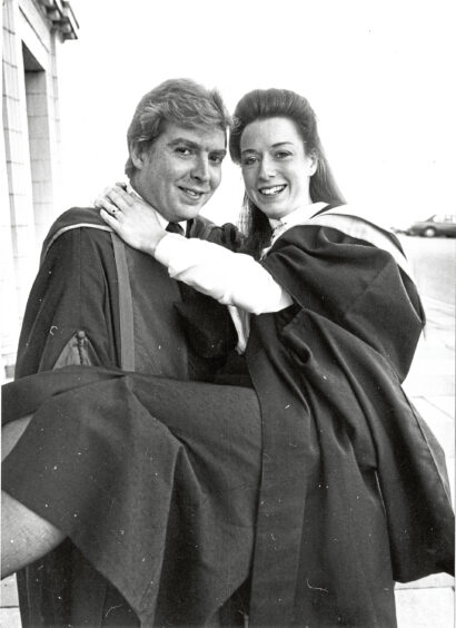 a woman in graduation robes being held in the arms of her fiance, who is wearing lecturer robes