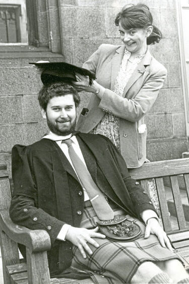 A woman places a graduation cap on a man sitting on a bench in traditional clothing