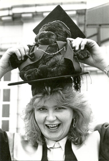 A woman in graduation robes balancing a stuffed monkey on her head, also wearing graduation robes