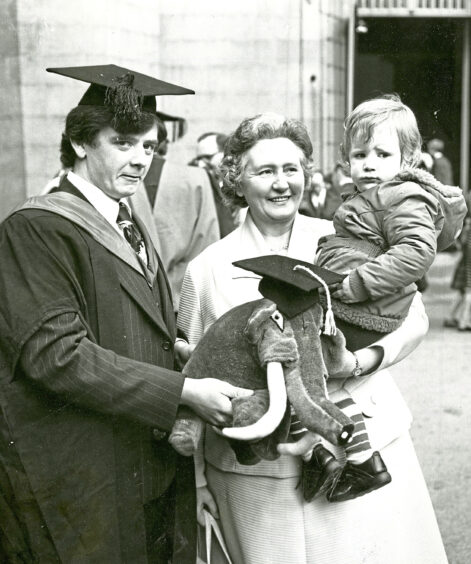 A man in gowns holding a stuffed elephant wearing a graduation cap and his mum holding a child