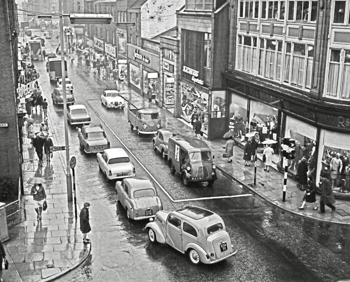 St Nicholas Street Aberdeen on a busy day- cars line the road and shoppers are walking on the pavement with umbrellas
