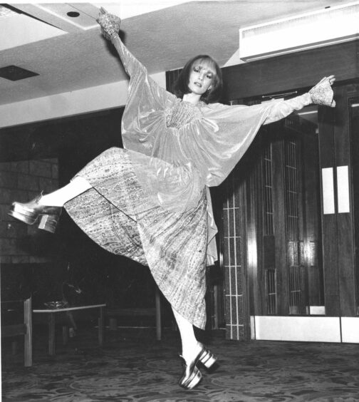 A model striking an exaggerated pose- her arms up in the air and a leg kicked out. She's wearing a draped blouse and a patterned wrapped skirt along with some platformed shoes
