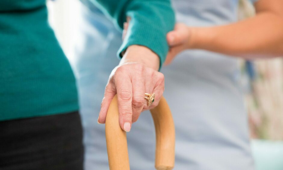 Old woman with hand on walking stick, with a care worker's hand holding her arm.