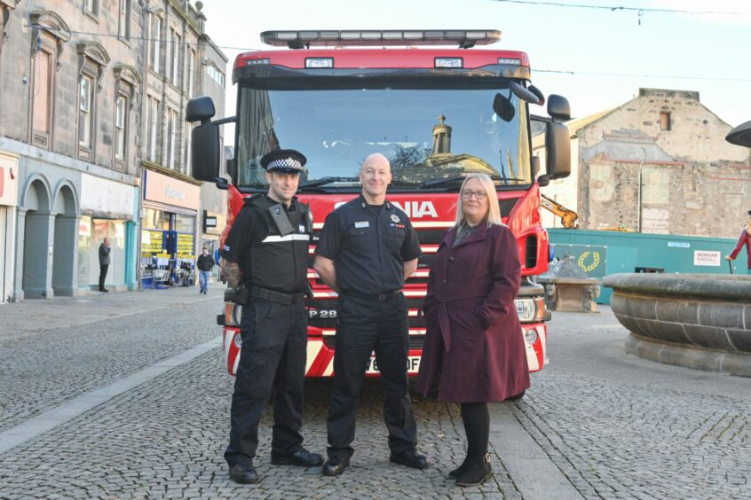 Jamie Dey Community Police Officer, Andy Munro Crew Commander and Elgin Bid manager Angela Norrie standing before a fire engine on the High Street in Elgin
