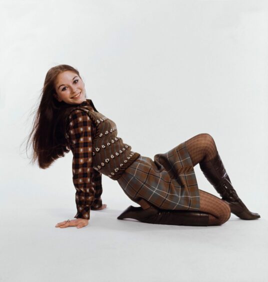 One of Bill Gibb's outfits on a young female model- the outfit consists of brown patterned pieces including a plaid skirt, checked shirt and jumper vest