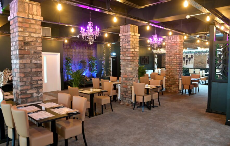 Mangrove's main sitting area, with sleek tables and comfortable looking chairs. Industrial-looking brick pillars stand throughout the room with neon lighting in chandeliers. 