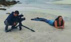 A man films a woman dressed as a mermaid, who is lying on a beach.
