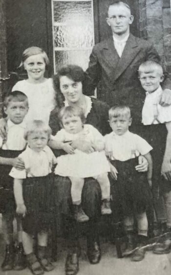A black and white image taken in Germany of Ilse Collinson and her parents, with siblings. She is standing behind her mother and next to her father as the oldest of the Quest children.