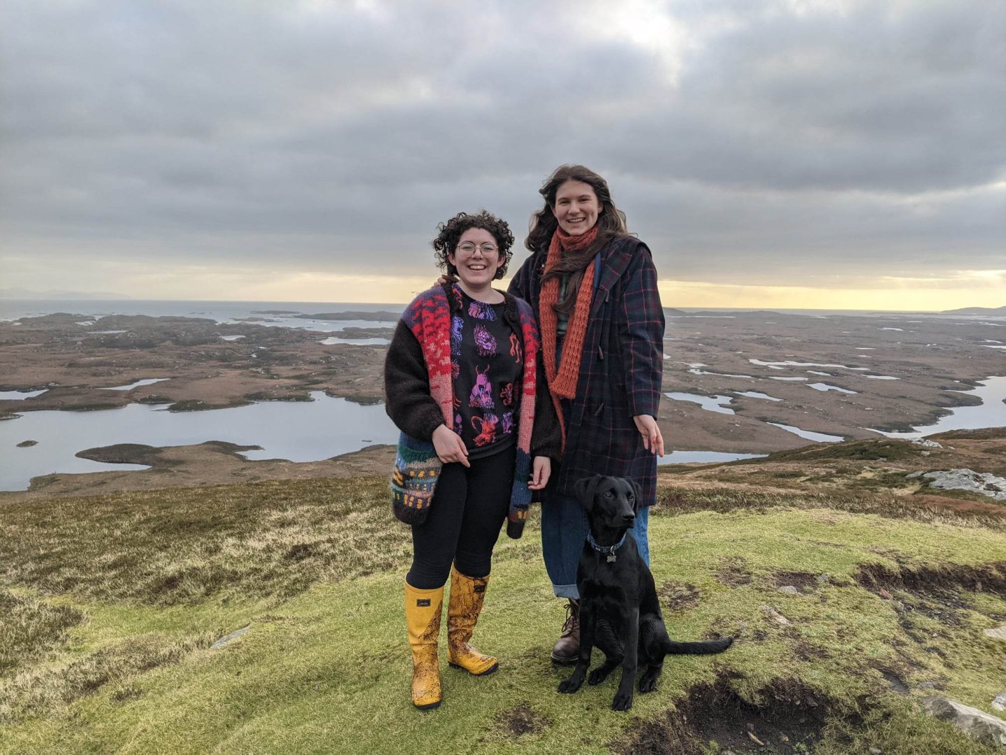 Two women and a dog stand on a windy hill.