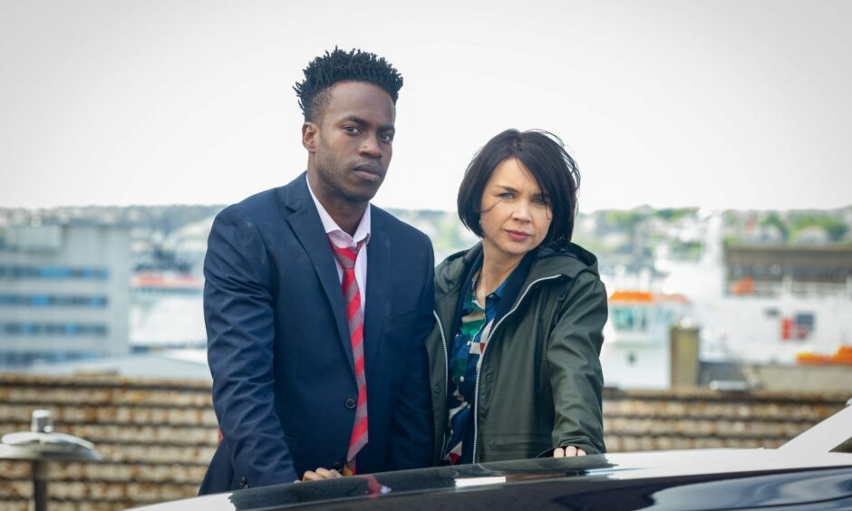 Actors Hannah Donaldson and Romario Simpson from Granite Harbour stare into the camera. Romario wears a dishevelled looking suit with Hannah to his right with windswept hair.