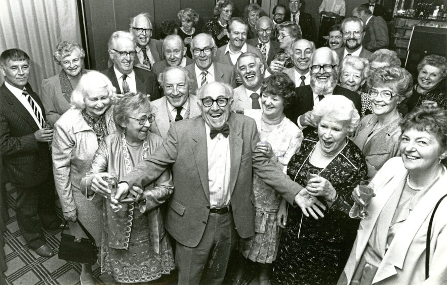 A crowd of elderly people holding champagne flutes smiling and laughing at the camera during a celebration
