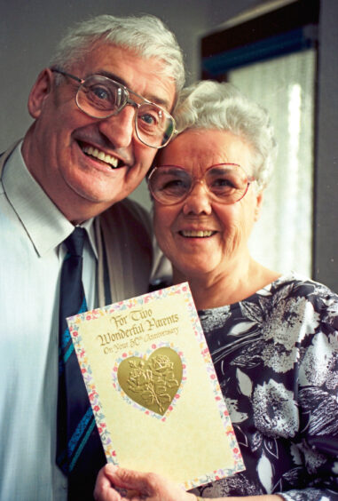 An elderly couple smiling at the camera with a golden wedding anniversary card in between them