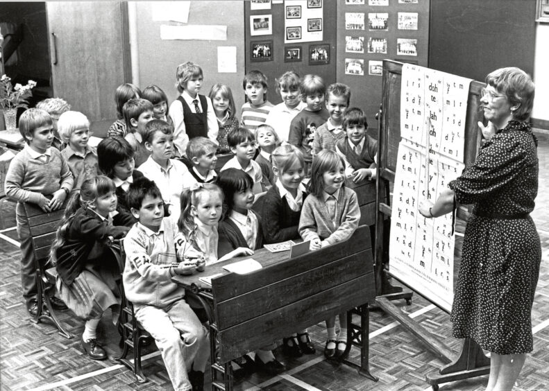 A teacher in front of a class of children pointing at a chart of words and letters on the anniversary of the school
