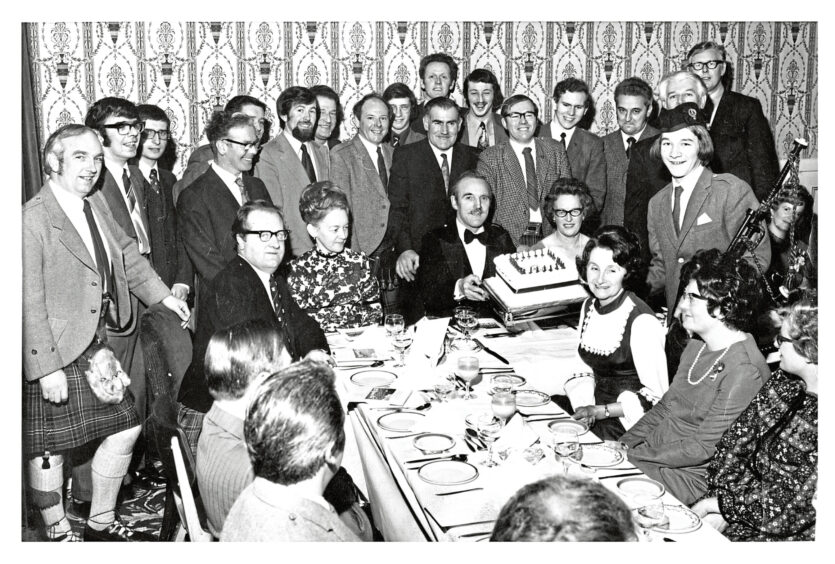 People crowded around a table in formal-wear, two people at the table are holding a large square cake