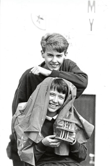 A girl with a flag draped over her and a game board in her hands is being leaned on by another pupil