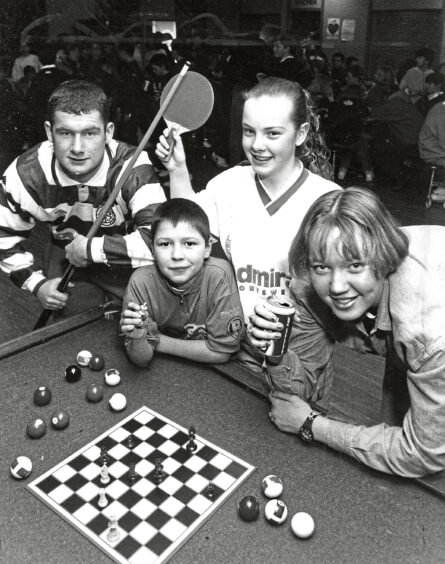 Four teenagers leaning on a pool table with a game of chess set up on it, the boy on the left is holding a pool cue, the boy next to him holds up a chess piece.. The two girls on the right are smiling at the camera, one holding a table tennis paddle and the other a can of juice