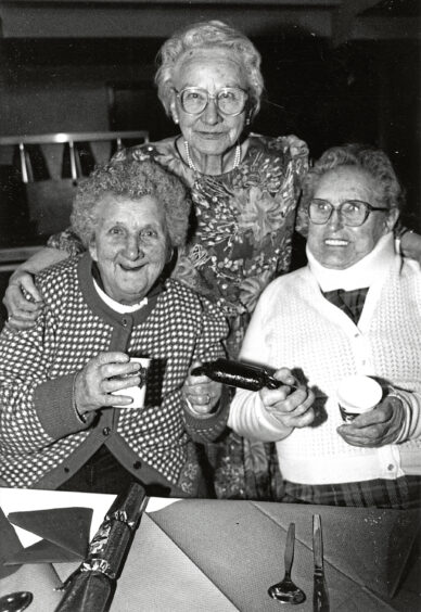 Three elderly women, two sitting down with a cracker between them and the other behind them with a hand on each shoulder