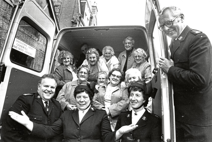 A group of elderly people smiling at the camera in the back of a minibus, behind four volunteers