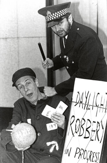 A man dressed as a policeman holds his baton above a man dressed as a prisoner next to a sign reading "daylight robbery in progress"