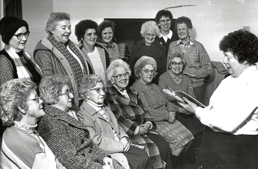 A group of elderly women sitting in two rows in front of a Salvation Army Aberdeen Major with a book