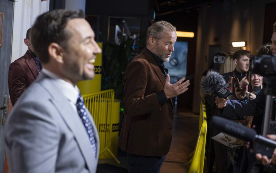 Iain Glen in focus gesturing on the red carpet of The Rig premiere with Martin Compston smiling, out of focus. 