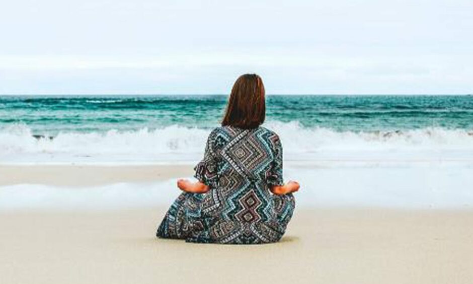 A woman sits with her back to the camera in a yoga pose on the beach.