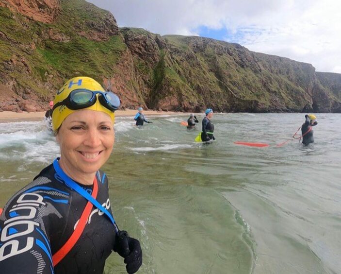A woman, wearing a wetsuit and swimming hat, takes a selfie showing the sea she is about to swim in.