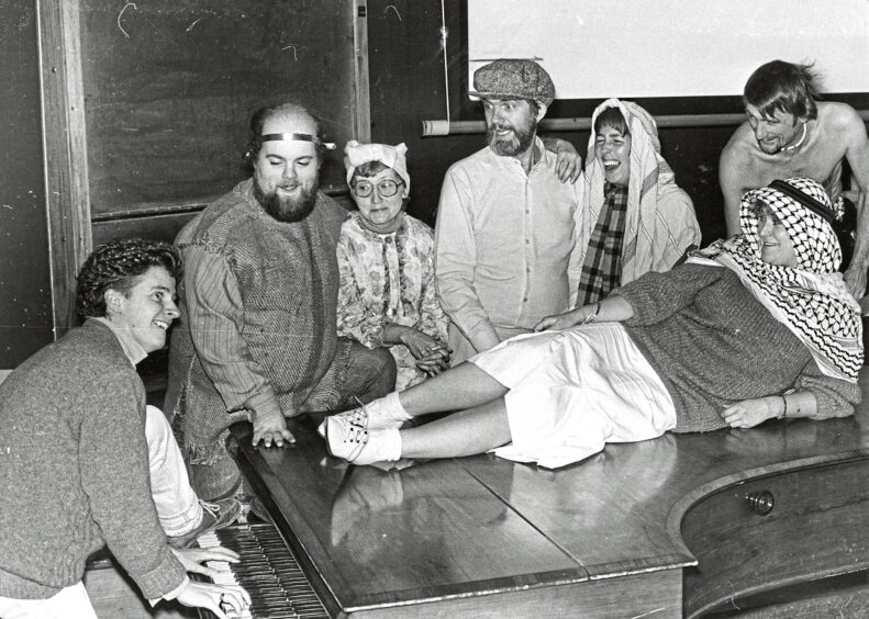A woman lying on top of a grand piano a man is playing with his foot up on the keys, surrounded by other costumed cast members