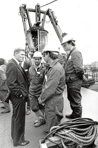 Men in safety gear speaking to a man in a suit with a large piece of machinery behind them