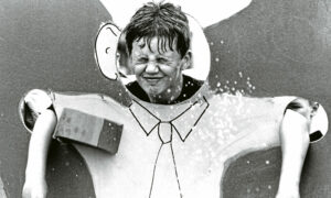 A boy with his head and arms poking through holes scrunches up his face as he is pelted with wet sponges