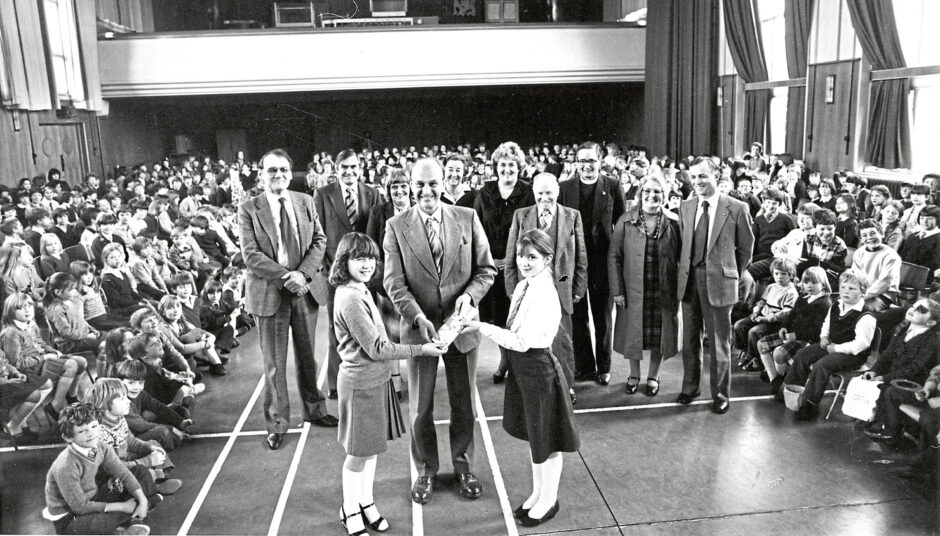 The assembly hall full of children surrounding, two pupils handing a man in a suit stamps