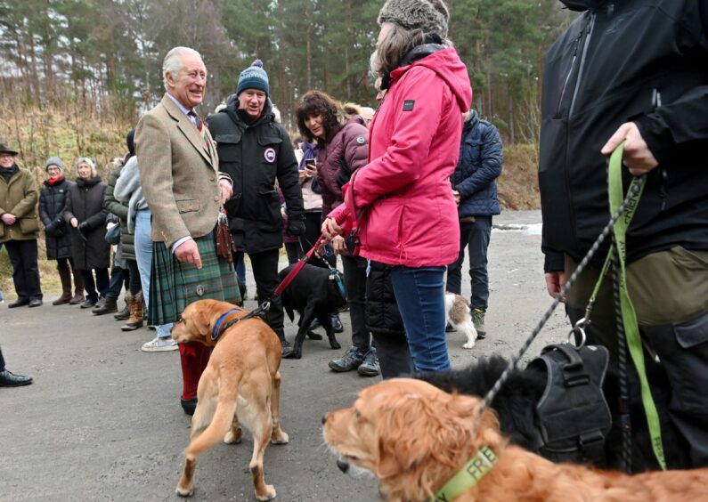 King Charles stands in front of members of the public in Aboyne. He is wearing a kilt and grinning while talking to a dog walker, whose dog appears to be sniffing the King. 