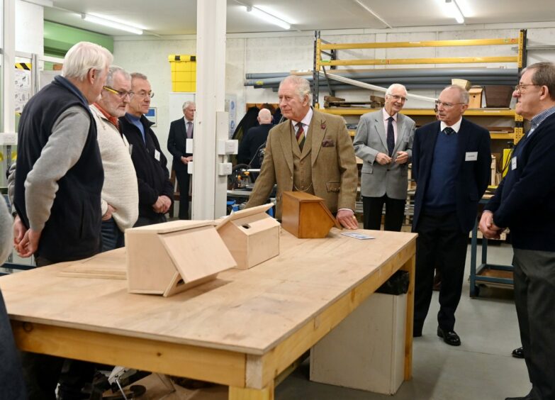 King Charles stands at the head of a wooden work bench, resting his hands on the table while in conversation with members of the Aboyne and Mid-Deeside Men's Shed.