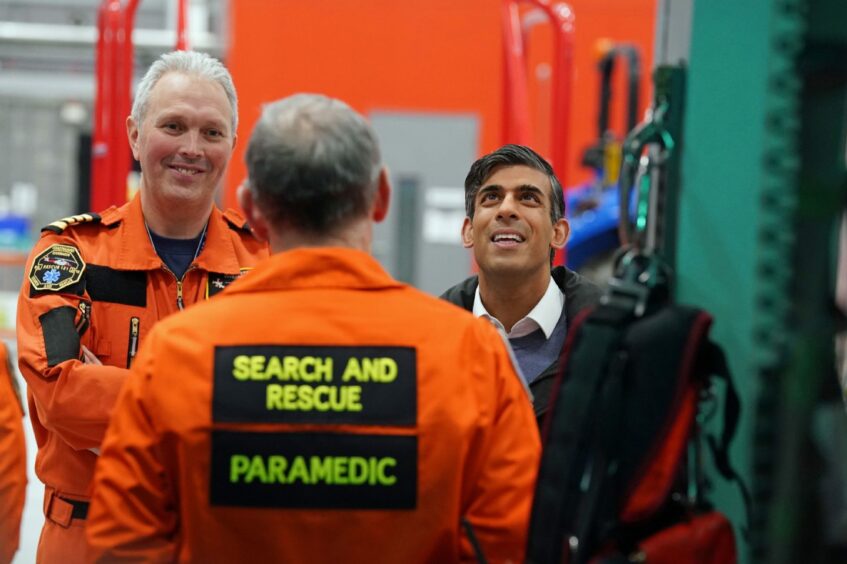 Rishi Sunak smiling and looking upward, with Captain Simon Hammock to his left smiling in his Coastguard uniform as they speak with a crew member during a visit to the Coastguard search and rescue base in Inverness Airport.
