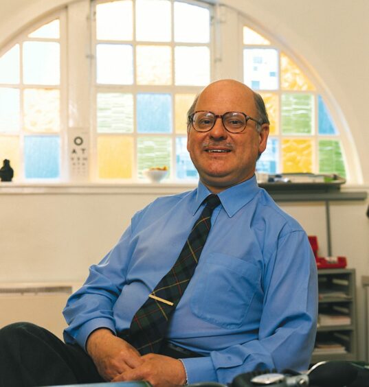 Shown during his time as a GP, Dr Roddy Macleod, originally from Skye. Pictured in a blue shirt and tartan tie sitting in his office with a large, bright arched window behind him.