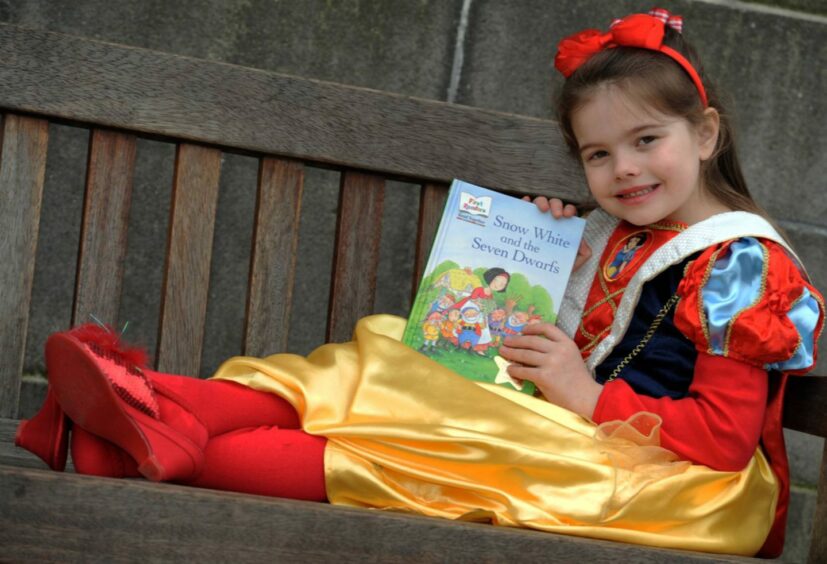 A young girl dressed as snow white sitting on a bench with her copy of snow white and the seven dwarves in her hands