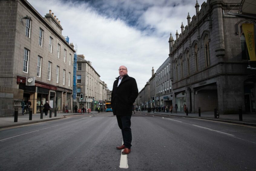 Bob Keiller, wearing a black coat, stands in the middle of Union Street with Jamieson & Carry and the Trinity Centre immediately behind him. He faces towards Union Terrace with the Castlegate behind him.