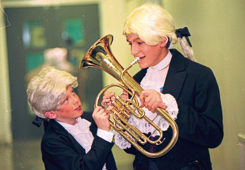 Two boys with cravats and wigs on holding a french horn.