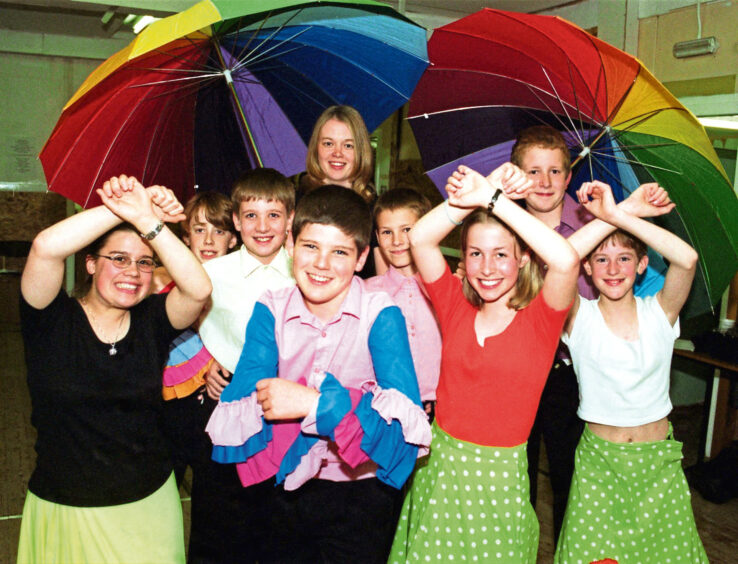Gang Show cast members in colourful costumes posing under two large rainbow coloured umbrellas.